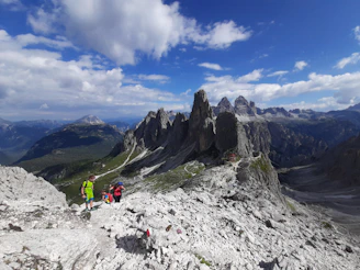 A rugged Himalayan mountain trail with trekkers crossing a narrow ridge under a clear blue sky.