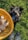 A small black dog is playfully reaching up towards a wicker basket filled with mushrooms. The setting is outdoors on green grass, and a pair of black, mud-splattered boots are visible at the bottom of the image.
