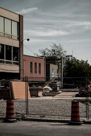 A construction site with a gravel ground and a fenced perimeter. In the background, there are multi-story buildings, with the facade showing windows and brickwork. There are construction materials and equipment scattered within the enclosed area. Two orange and black striped traffic barrels stand in front of the fence, and trees are visible in the background under a cloudy sky.