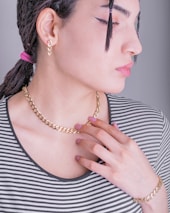 A person with a side profile pose, wearing a striped shirt and accessorized with a gold chain necklace, a matching bracelet, and a pair of earrings. The hair is styled in a braid with a pink hair tie, and there is noticeable makeup with eyeliner. The person’s hand is delicately touching the necklace.