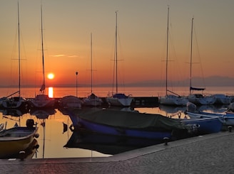 Calm harbor at sunset with colorful boats anchored and soft orange light reflecting on water.