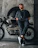 Close-up of a biker wearing a dark Stillroll hoodie, leaning against a vintage motorcycle under moody lighting.