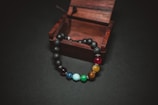 A multicolored bead bracelet is partially placed inside an open, small wooden box. The beads are made of various stones, each showcasing distinct colors, including black, red, yellow, green, blue, and white. The backdrop is a dark surface, which contrasts with the vibrant colors of the beads.