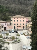 A picturesque town square with a central fountain surrounded by benches and young trees. The area is lined with historic buildings painted in soft pink tones with green shutters. Several cars are parked along the street, and a few people are walking around. The backdrop consists of dense, leafless trees suggestive of winter or early spring.