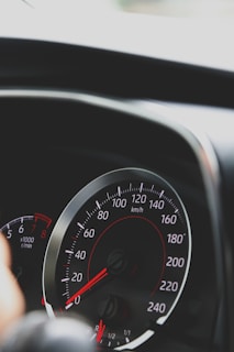 A close-up view of a car speedometer with red and white markings measuring speed in kilometers per hour. A portion of the dashboard is visible, creating a dynamic and sleek look.