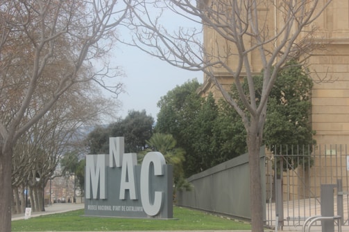 Large metallic letters displaying the acronym 'MNAC' are positioned near bare trees and a section of a classical building. The scene is set in an outdoor environment with a pathway leading alongside the building.