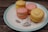 A plate featuring four colorful macarons arranged neatly. The macarons are a mix of pink and yellow hues with creamy fillings, placed on an ornate plate adorned with floral patterns. The setting appears rustic with a wooden backdrop providing a textured contrast.