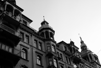 An architectural shot depicting a series of classic buildings with intricate design elements. The structures exhibit ornate balconies, detailed window frames, and a range of roof shapes. The image is captured in black and white, highlighting the textures and lines of the facades under a clear sky.