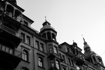 An architectural shot depicting a series of classic buildings with intricate design elements. The structures exhibit ornate balconies, detailed window frames, and a range of roof shapes. The image is captured in black and white, highlighting the textures and lines of the facades under a clear sky.