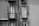 Two adjacent windows on a building facade feature balconies with ornate wrought-iron railings. Each window has closed shutters, and the building wall shows textures that indicate a vintage or aged exterior. The shadow of the railings creates intricate patterns on the wall.