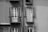 Two adjacent windows on a building facade feature balconies with ornate wrought-iron railings. Each window has closed shutters, and the building wall shows textures that indicate a vintage or aged exterior. The shadow of the railings creates intricate patterns on the wall.