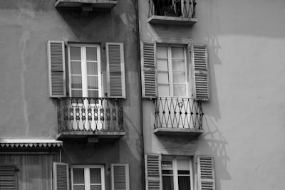 Two adjacent windows on a building facade feature balconies with ornate wrought-iron railings. Each window has closed shutters, and the building wall shows textures that indicate a vintage or aged exterior. The shadow of the railings creates intricate patterns on the wall.