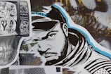 A stylized street art collage features a monochrome illustration of a man’s face wearing a hood. The artwork incorporates bold black and white lines with blue accents. To the left, a black and white print displaying cartoon characters overlaid on a TV screen adds a nostalgic element. A variety of textures and abstract backgrounds are present, contributing to a dynamic urban aesthetic.