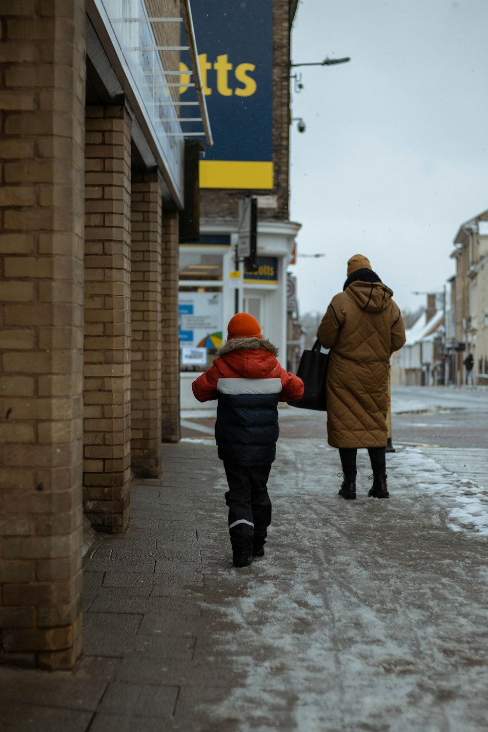 woman in brown coat and black pants walking on snow covered road during daytime