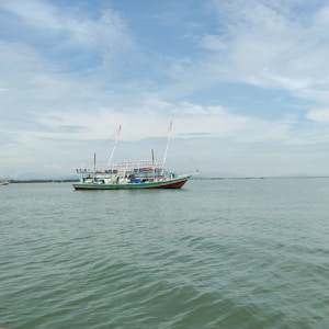 A fishing boat with a colorful hull is floating on calm waters under a lightly clouded sky. The boat has tall masts and equipment on deck, positioned in a tranquil seascape setting.