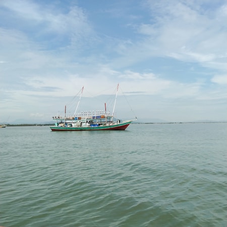 A fishing boat with a colorful hull is floating on calm waters under a lightly clouded sky. The boat has tall masts and equipment on deck, positioned in a tranquil seascape setting.