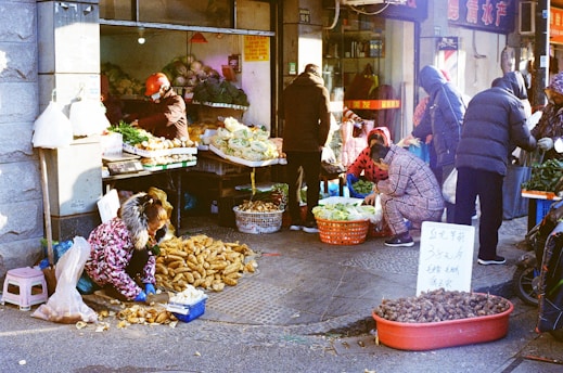 A bustling outdoor market scene where several people are engaged in buying and selling vegetables. Various produce such as potatoes and leafy greens are displayed in baskets and on tables. The vendors and customers are bundled up in winter clothing, suggesting cold weather. There is a handwritten sign with prices displayed among the goods.