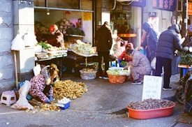 A bustling outdoor market scene where several people are engaged in buying and selling vegetables. Various produce such as potatoes and leafy greens are displayed in baskets and on tables. The vendors and customers are bundled up in winter clothing, suggesting cold weather. There is a handwritten sign with prices displayed among the goods.