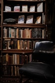 A dimly lit bookshelf filled with a variety of books, some of which are positioned upright while others are leaning. The shelves also display a vintage typewriter and a few framed book covers. In front of the bookshelf is a dark, worn-out armchair, suggesting a cozy reading nook.