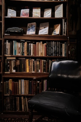 A dimly lit bookshelf filled with a variety of books, some of which are positioned upright while others are leaning. The shelves also display a vintage typewriter and a few framed book covers. In front of the bookshelf is a dark, worn-out armchair, suggesting a cozy reading nook.