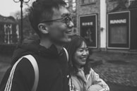 A candid shot of two friends laughing, one in a soft beige trench coat, the other in a tailored black jacket, walking through a sunlit city street.