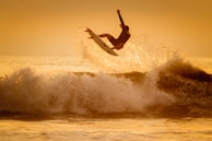 A surfer is performing an aerial maneuver above a wave during sunset. The scene is bathed in warm golden tones, highlighting the dynamic motion of the surfer and the spray of the ocean.