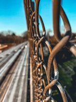 A close-up view of a chain-link fence with a blurred background. The metal of the fence appears rusted and weathered, providing a textured foreground. In the distance, elements like a road or pathway under bright blue skies are visible, with leafless trees indicative of late fall or winter.