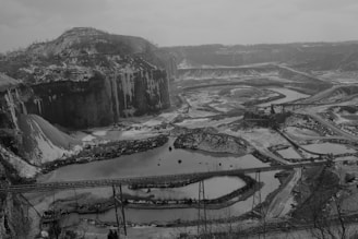 A vast quarry landscape with layered rock formations and large open pits. There are multiple ponds of water with winding roads and pathways cutting through the terrain. The area is covered in snow, adding a stark contrast to the dark rock surfaces.
