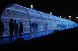 A cylindrical tunnel covered in blue LED lights, creating a glowing path. Several people stand inside or near the tunnel, some facing one another and engaging in conversation. The tunnel provides a futuristic and luminous ambiance, standing out against the dark night sky. In the background, some tall buildings with illuminated signs are visible.