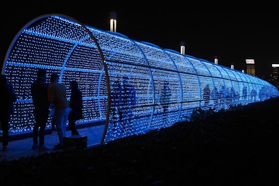 A cylindrical tunnel covered in blue LED lights, creating a glowing path. Several people stand inside or near the tunnel, some facing one another and engaging in conversation. The tunnel provides a futuristic and luminous ambiance, standing out against the dark night sky. In the background, some tall buildings with illuminated signs are visible.