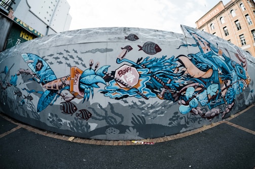 A dynamic and colorful mural depicting an underwater scene with stylized marine life. The artwork features a variety of sea creatures, including fish and crabs, intertwined with abstract blue and orange shapes. The background is filled with silhouettes of fish, giving the impression of a bustling underwater world.