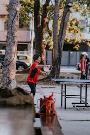 A person is actively engaged in a game of table tennis outdoors. The setting is a park-like area with tall trees providing shade. In the background, there are parked cars and a house with a tiled roof. The player is mid-action, preparing to hit the ball with a paddle.