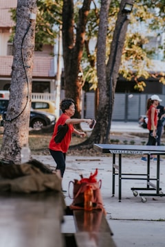 A person is actively engaged in a game of table tennis outdoors. The setting is a park-like area with tall trees providing shade. In the background, there are parked cars and a house with a tiled roof. The player is mid-action, preparing to hit the ball with a paddle.