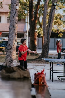 An elderly person wearing a red shirt is engaged in a game of table tennis in an outdoor setting surrounded by trees. Another person can be seen in the background, also dressed in a red shirt, possibly waiting for their turn. A table tennis table with the brand name 'Challenger' is in front of the player. Nearby, there are items placed on a bench, including a brown bag and a bright orange water bottle.