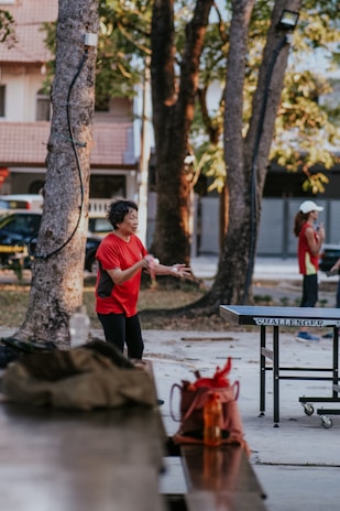 An elderly person wearing a red shirt is engaged in a game of table tennis in an outdoor setting surrounded by trees. Another person can be seen in the background, also dressed in a red shirt, possibly waiting for their turn. A table tennis table with the brand name 'Challenger' is in front of the player. Nearby, there are items placed on a bench, including a brown bag and a bright orange water bottle.