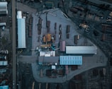 An aerial view of an industrial area with multiple buildings, storage areas, and stacks of logs or lumber. The layout includes long rectangular structures, open spaces with arranged materials, and various equipment positioned around the site.