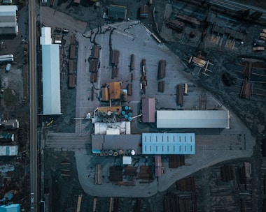 An aerial view of an industrial area with multiple buildings, storage areas, and stacks of logs or lumber. The layout includes long rectangular structures, open spaces with arranged materials, and various equipment positioned around the site.