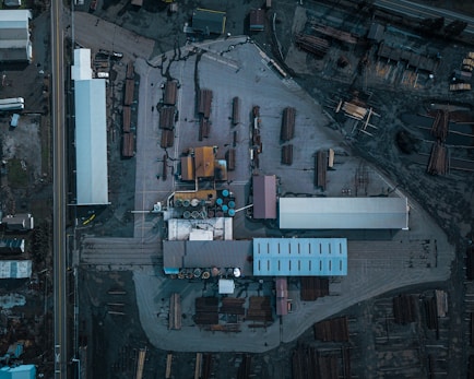 An aerial view of an industrial area with multiple buildings, storage areas, and stacks of logs or lumber. The layout includes long rectangular structures, open spaces with arranged materials, and various equipment positioned around the site.