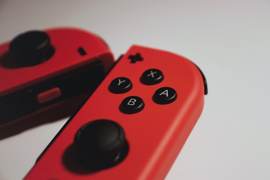 Two red gaming controllers with button labels Y, X, A, and B are positioned on a light surface. The controllers have black thumbsticks and buttons.