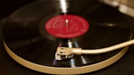 A close-up view of a vinyl record being played on a turntable. The record is spinning, and the tonearm is positioned over it. The center label of the record is red, contrasting with the black vinyl.
