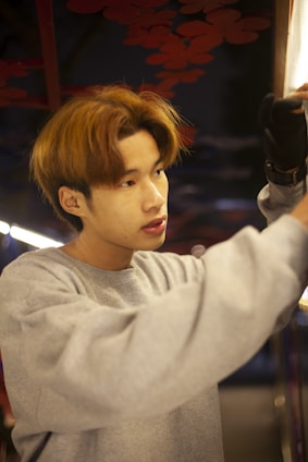 A person wearing a grey sweatshirt and gloves appears to be focused on an activity using their hands, possibly adjusting or fixing something. The background features a dark setting with red leaves or decorative elements visible.