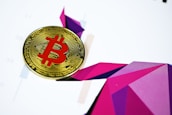 A Bitcoin coin with a red symbol is placed on a colorful geometric abstract background. The background features various shades of purple and pink geometric shapes, creating a dynamic and modern visual effect.