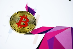 A Bitcoin coin with a red symbol is placed on a colorful geometric abstract background. The background features various shades of purple and pink geometric shapes, creating a dynamic and modern visual effect.