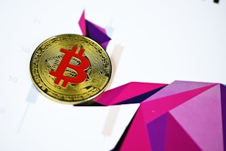 A Bitcoin coin with a red symbol is placed on a colorful geometric abstract background. The background features various shades of purple and pink geometric shapes, creating a dynamic and modern visual effect.