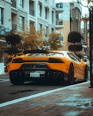 A sleek yellow sports car is parked along a street in an urban area with modern buildings in the background. The car is a Lamborghini, identified by its distinctive design and visible logo. Greenery and shop signs are present, adding to the cityscape atmosphere.