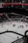 An ice hockey game is in progress in a large indoor arena. Players wearing uniforms in red and white are engaged in play near one of the goals. The stadium seating is dimly lit, and the scoreboard displays a score with red numerical indicators. A referee, dressed in black and white stripes, stands on the ice.