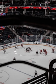 An ice hockey game is in progress in a large indoor arena. Players wearing uniforms in red and white are engaged in play near one of the goals. The stadium seating is dimly lit, and the scoreboard displays a score with red numerical indicators. A referee, dressed in black and white stripes, stands on the ice.