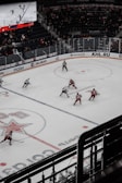 A hockey game taking place in an indoor arena with players in red and white uniforms competing on an ice rink. The arena has spectators in the stands, and a large digital screen displays the game. The rink is marked with various lines and logos.