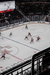A hockey game taking place in an indoor arena with players in red and white uniforms competing on an ice rink. The arena has spectators in the stands, and a large digital screen displays the game. The rink is marked with various lines and logos.