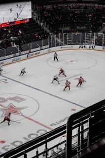 A hockey game taking place in an indoor arena with players in red and white uniforms competing on an ice rink. The arena has spectators in the stands, and a large digital screen displays the game. The rink is marked with various lines and logos.